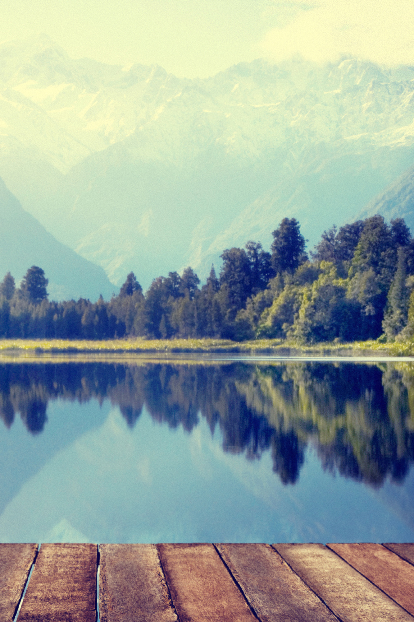 Landscape photograph taken from a dock overlooking a calm lake with trees and mountains and clouds in the background, which are reflected off of the water like a mirror. 